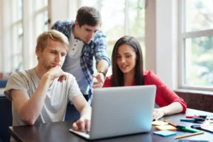 Three individuals collaborating on a laptop in a modern office setting, focused on an AI and ML course project.